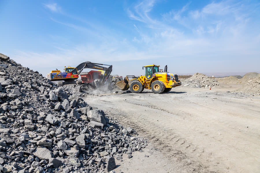 MySol excavator and dump truck moving bulk material at the foot of a large stockpile.